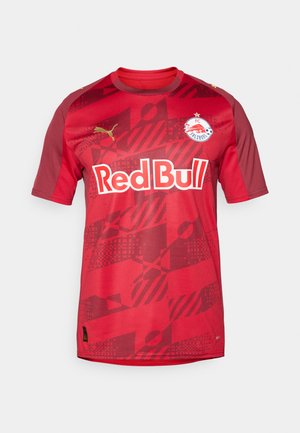 Camiseta de fútbol roja con mangas cortas, cuello redondo y un patrón degradado. En la parte frontal se muestra el logo "Red Bull", el escudo del equipo y el logo dorado de Puma.