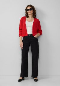 Rode gebreide cardigan over een witte tanktop, gecombineerd met zwarte high-waisted jeans en zwarte platte schoenen. Model draagt zonnebril.