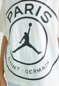 Un t-shirt en coton blanc avec un grand graphisme noir représentant une silhouette sautant et le texte "PARIS" en haut, "SAINT - GERMAIN" en bas dans un design circulaire.