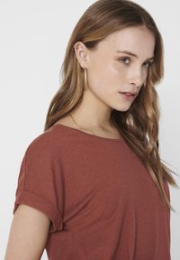 T-shirt color ruggine con maniche arrotolate, morbido al tatto, vestibilità rilassata e scollo rotondo, indossato da una persona con capelli mossi.