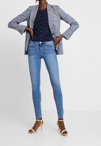 Blazer de cuadros gris sobre una blusa azul marino con botones, combinado con jeans ajustados azul claro y tacones amarillos con tiras. Diseño simple y ajustado.