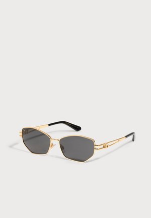 Lunettes de soleil dorées aux verres anguleux gris foncé, avec des plaquettes nasales transparentes et des branches terminées par des embouts noirs, sur fond clair.