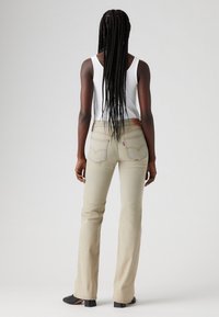 Jeans beige claro acampanados con una cintura de media altura, bolsillos traseros y costuras contrastantes. Combinados con una camiseta sin mangas blanca y calzado negro.
