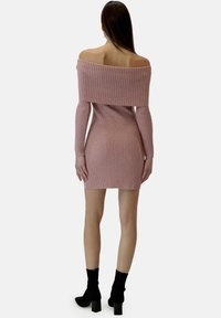 Schulterfreies, gerippt gestricktes Kleid in sanftem Rosa, figurbetonte Silhouette, lange Ärmel und Mini-Länge. Kombiniert mit schwarzen Stiefeletten mit Absatz.