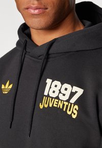 Svart hoodie i bomull med vit och gul "1897 JUVENTUS" tryck och en liten gul Adidas-logga på övre vänstra sidan.