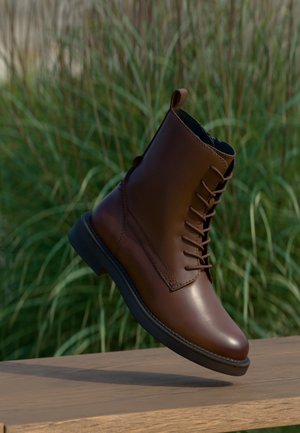 Botte marron en cuir à lacets avec semelle noire flottant au-dessus d'une surface en bois avec un fond d'herbe verte.