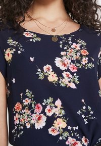 Haut floral bleu marine à manches courtes, avec des fleurs roses claires et rouges, des feuilles vertes et une texture douce et lisse. Superposition de délicates colliers en or.