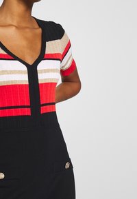 La robe côtelée possède un décolleté en V, des manches courtes et un design en block de couleurs rouge, blanc, or et noir, avec des boutons décoratifs en or.