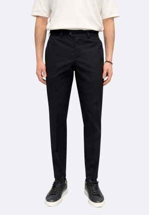 Uomo che indossa pantaloni neri aderenti, scarpe da ginnastica nere con suole bianche e una camicia bianca a maniche corte, in piedi dritto su uno sfondo neutro.