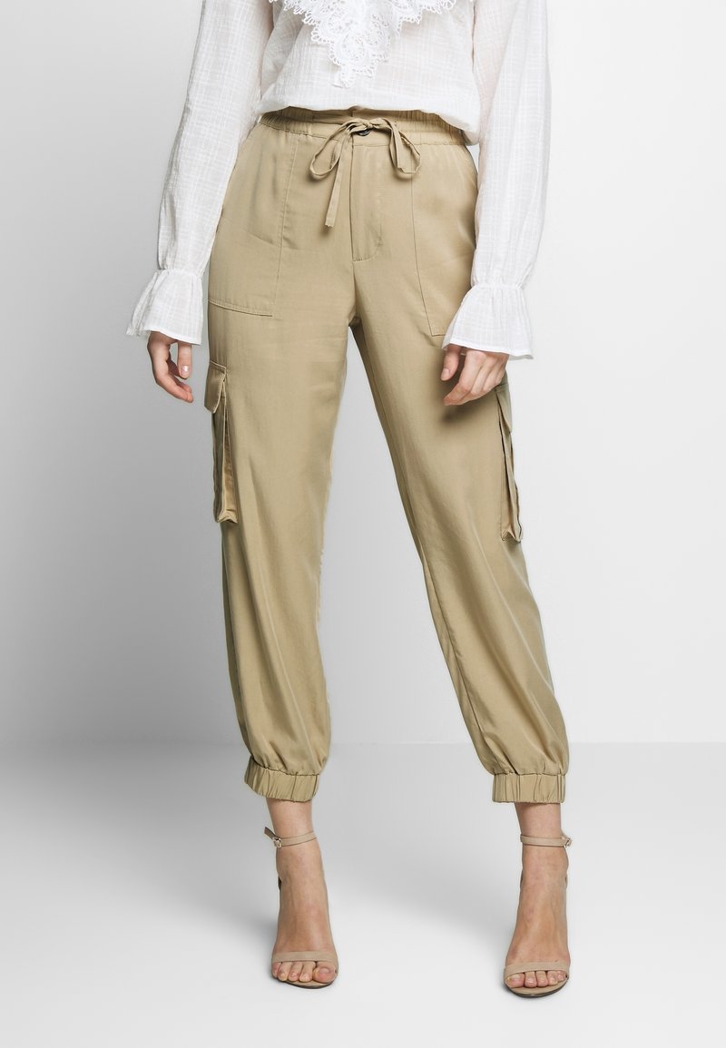 Pantalon cargo beige avec taille à cordon, poignets élastiques, poches latérales et tissu lisse. Porté avec un chemisier blanc et des sandales à bride cheville.