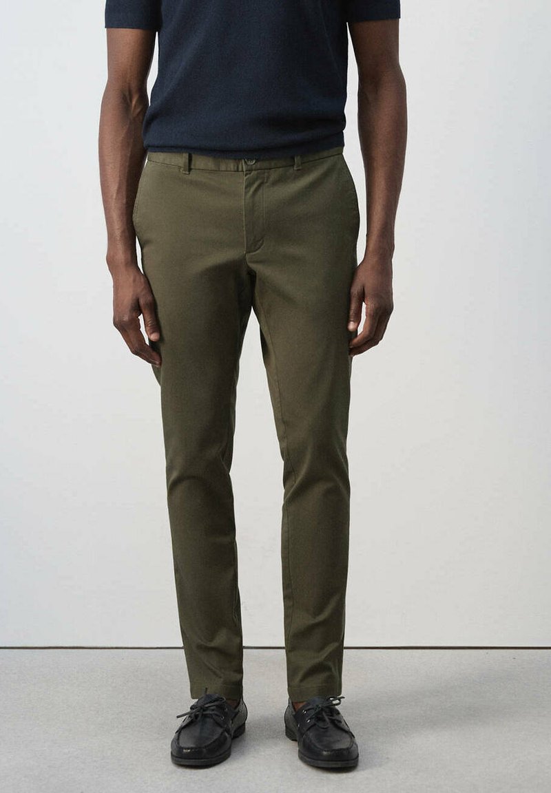 Homme portant un pantalon slim vert olive, des chaussures noires et une chemise sombre à manches courtes, debout, les bras détendus le long du corps.