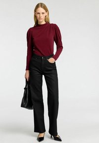 Bordeaux långärmad topp med puffiga axlar, kombinerad med högmidjade svarta jeans och spetsiga svarta klackar. Håller en svart väska.