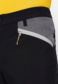 Czarne spodnie z szarą kieszenią z tkaniny, z białym suwakiem i żółtą zawieszką. Materiał jest gładki, zaprojektowany z myślą o funkcjonalności.