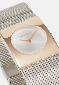 Montre avec un bracelet en maille argentée, un boîtier rectangulaire en doré, un cadran circulaire blanc et des poignées des heures et des minutes en or minimalistes.