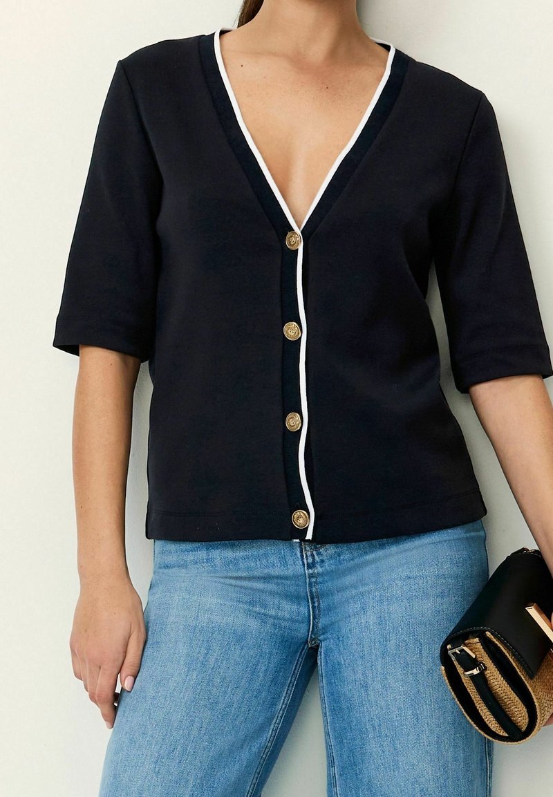 Navy short-sleeve cardigan with a V-neck, white trim, and golden buttons. Worn with light blue jeans and a black handbag.