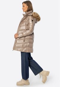 Mujer con abrigo acolchado beige con capucha con borde de piel, vaqueros de pierna ancha azul oscuro y botas beige gruesas con cordones, de pie de perfil.