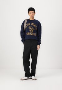 Sweatshirt azul-marinho com texto e gráfico em dourado, combinado com calças pretas e ténis brancos. Modelo leva uma mala cinza à tiracolo.
