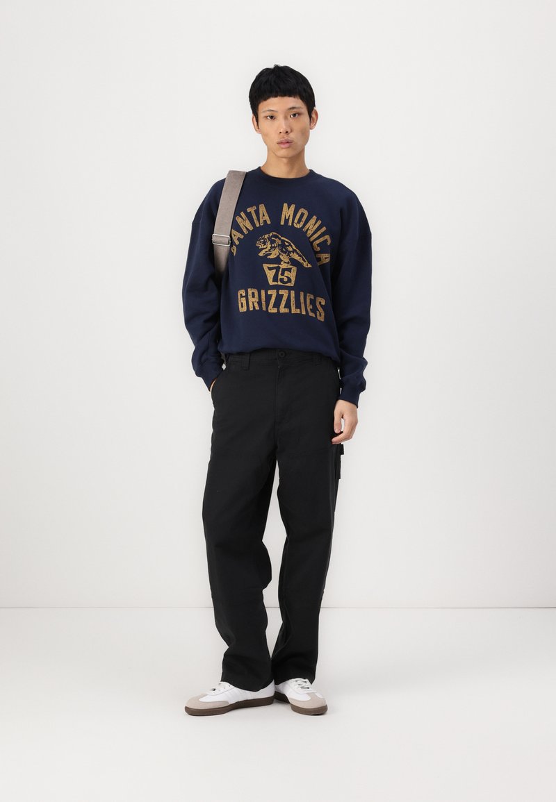 Sweatshirt navy con testo e grafica dorati, abbinato a pantaloni neri e sneakers bianche. Il modello porta una borsa a tracolla grigia.