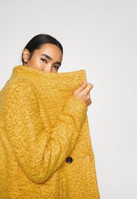 Cappotto di lana giallo senape con una finitura testurizzata, caratterizzato da un colletto alto e un bottone singolo. L'avvolgimento del tessuto oscura parzialmente il volto.