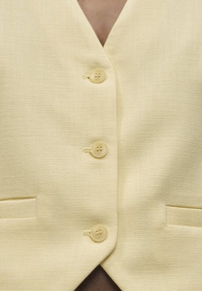 Κοντινή λήψη ενός γιλέκου από χλωμό κίτρινο ύφασμα με υφή, με τρία στρογγυλά κουμπιά και δύο οριζόντιες τσέπες με σχισμές.