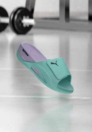 Een turquoise en lavendelkleurige Puma Nitro slippersandaal met open teen en verstelbare band, fitnessgewichten wazig op de achtergrond.