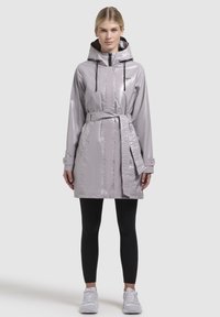 Pelerină de ploaie gri deschis, lucioasă, cu glugă, șnururi reglabile, talie cu brâu și două buzunare laterale. Purtați peste leggings negri și adidași.