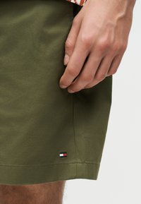 Κοντινή λήψη ενός χεριού που ακουμπά δίπλα σε χακί πράσινα σορτς με ένα μικρό ορθογώνιο κόκκινο, άσπρο και μπλε λογότυπο κοντά στο τελείωμα.