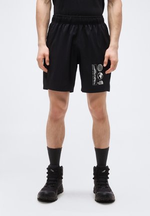 Homme portant un short de sport noir avec le logo et le texte blanc The North Face sur la jambe droite, accompagné de chaussettes noires et de chaussures de randonnée.