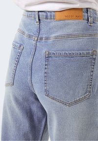 Ljust blå denimjeans med hög midja, synliga bakfickor och en brun läderetikett med texten "NOISY MAY" på midjebandet.