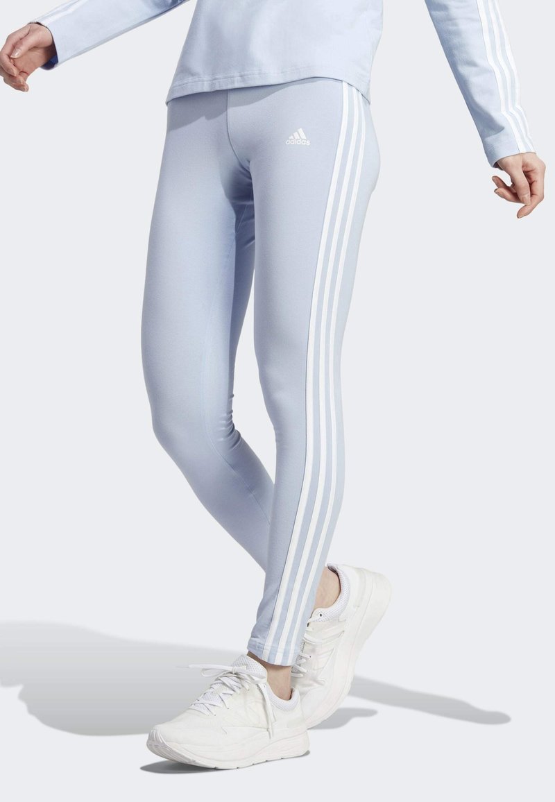 Persona vistiendo leggings de Adidas azul claro con rayas blancas y zapatillas blancas, de pie con una pierna cruzada sobre la otra.