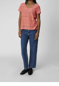 Blouse courte à manches courtes couleur corail rose avec motif à œillets et décolleté en V, associée à un jean en denim bleu à jambes larges et des ballerines noires.
