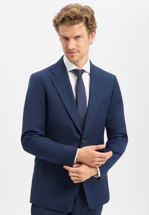 Man in navy blue suit and tie adjusting left sleeve with a focused expression, standing against a plain white background.