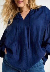 Marinblå, lätt vikt, tröja med v-ringning, framveck och dragsko. Långa dolmanärmar med avslappnad passform. Avslappnad design.