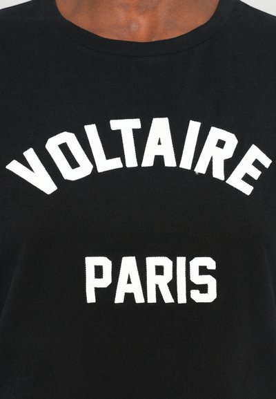Melnā kokvilnas T-kreklā ir uzraksts "VOLTAIRE" ar baltu lielu fontu, kas ir izliektā formā, un zem tā ir teksts "PARIS", kā arī klasiskā apaļā kakla dizains.