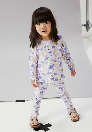 Jeune fille debout sur un X en ruban noir, portant un pyjama pastel à imprimé Hello Kitty et des sandales roses, sur un fond clair uni.