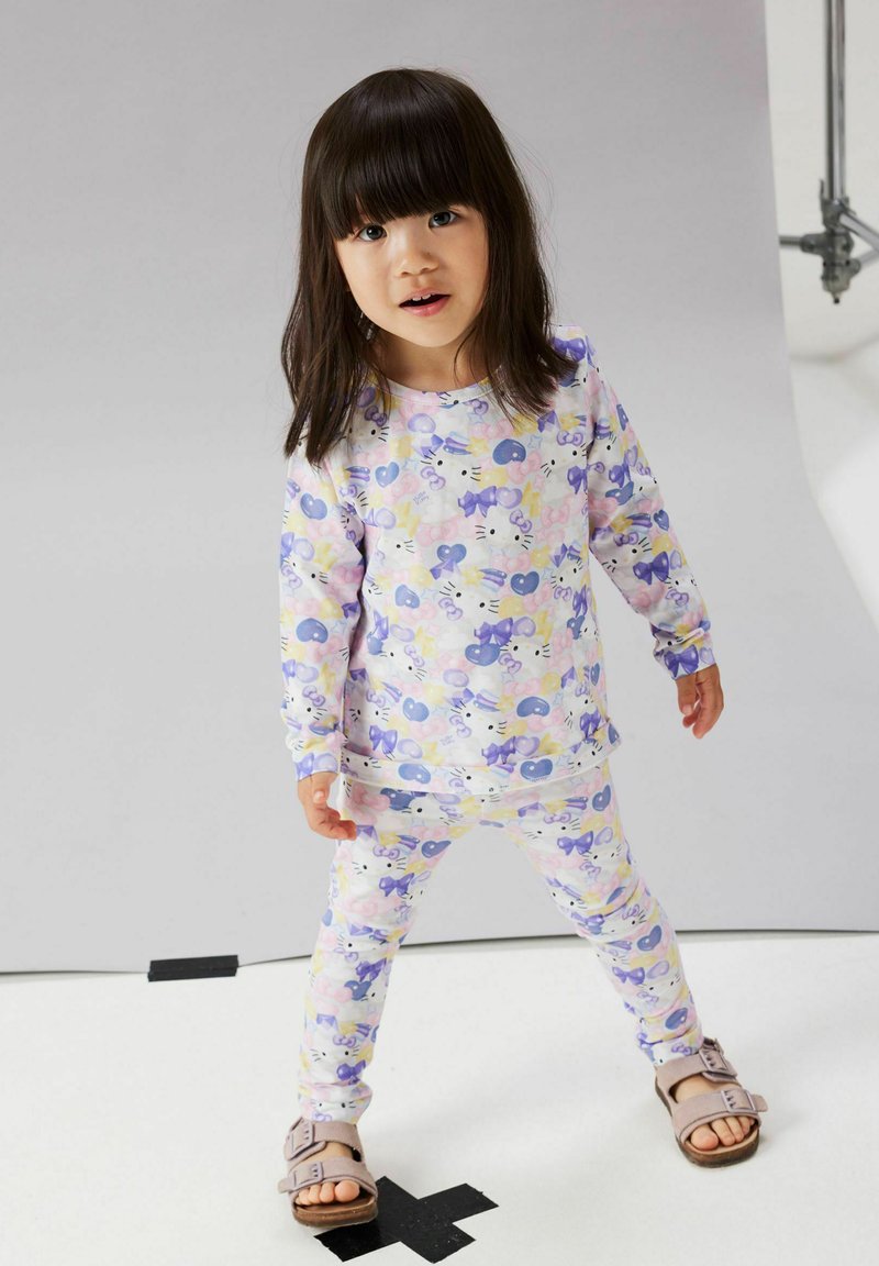 Jeune fille debout sur un X en ruban noir, portant un pyjama pastel à imprimé Hello Kitty et des sandales roses, sur un fond clair uni.