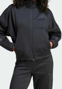Svart zip-up hoodie i mjukt tyg, med en framficka, avslappnad passform och tonala logotypdetaljer. Matchas med svarta joggers.