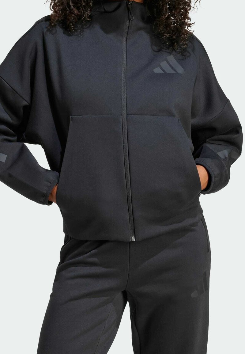 Svart zip-up hoodie i mjukt tyg, med en framficka, avslappnad passform och tonala logotypdetaljer. Matchas med svarta joggers.