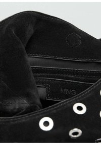 Interior din piele întoarsă neagră al unei încălțări, cu inele metalice argintii și o etichetă de material textil cu textul "MNG." Textură moale și margini rotunjite vizibile.