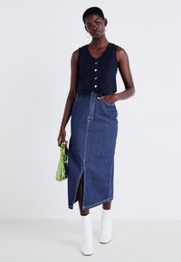 Τζιν φούστα με μπροστινή σχισμή, συνδυασμένη με ναυτικό μπλε crop top με κουμπιά. Το μοντέλο κρατάει μία πράσινη, διαφανή τσάντα και φοράει λευκές μπότες μέχρι τον αστράγαλο.