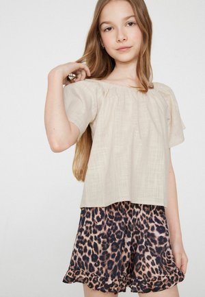 Joven chica con cabello largo y castaño, con una blusa beige sin hombros y pantalones cortos con estampado de leopardo y dobladillos con volantes sobre un fondo liso.