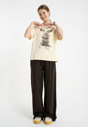 Uśmiechnięta młoda kobieta z kucykiem nosi beżowy T-shirt z króliczkiem i napisem "Free Hugs", czarne spodnie o szerokich nogawkach oraz beżowe buty na platformie.