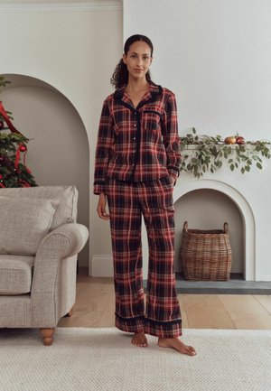 Mujer de pie descalza con pijama de cuadros rojos y negros en una sala de estar acogedora con un árbol de Navidad y una chimenea con vegetación y adornos.