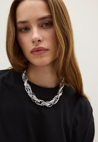 Chunky zilveren ketting met in elkaar verbonden metalen lussen, gedragen over een zwart sweatshirt. Glad, reflecterend oppervlak met een gedurfde uitstraling.