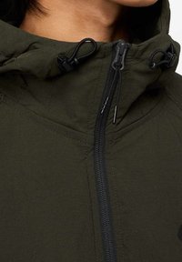 Olivgrön jacka med hög krage, synlig svart dragkedja, justerbara snörningar och en slät yta. Har förstärkta sömdetaljer.