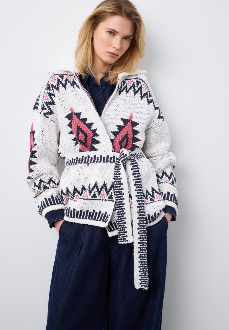 Frau trägt weiße Strickjacke mit roten und schwarzen geometrischen Mustern, die an der Taille gebunden ist, kombiniert mit dunkelblauer Hose und Hemd.