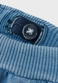 Tessuto blu chiaro con texture a righe e una vita blu scuro. Presenta un bottone trasparente con un design floreale e cuciture in colori a contrasto.