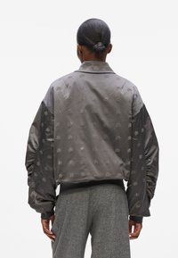 Giacca bomber grigia con una finitura satinata lucida, modelli ricamati e maniche voluminose. I polsini e l'orlo a coste completano il design.