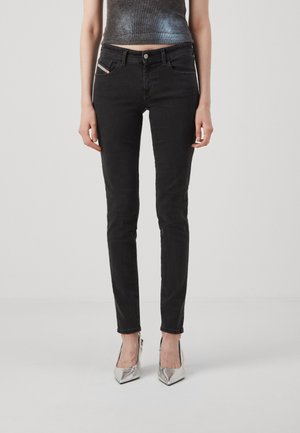 Jean noir slim porté avec des talons aiguilles argentés pointus et un top gris court sans manches sur un fond uni.