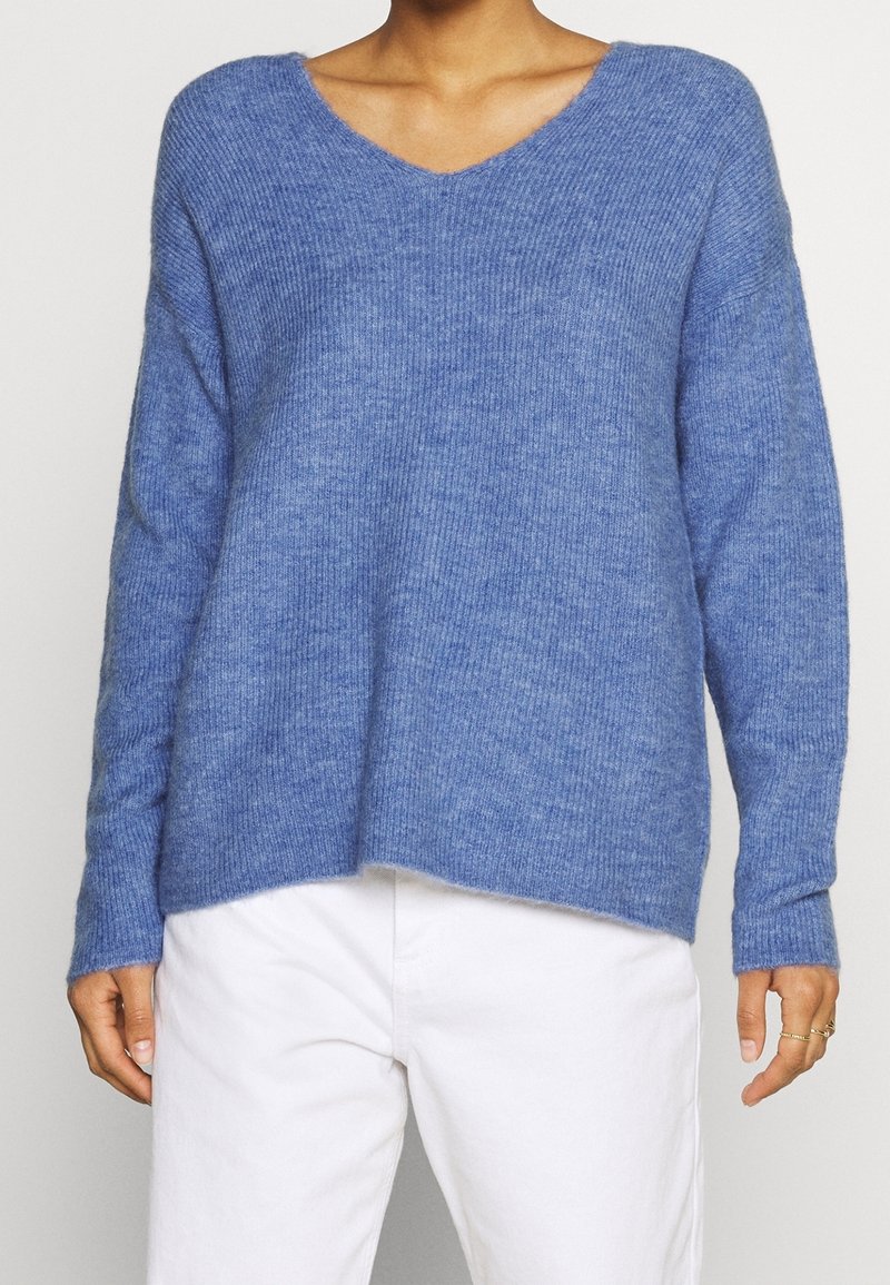 Femme portant un pull en maille bleu à manches longues avec un col en V et un pantalon blanc, debout devant un fond uni.